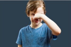A young child in a blue t-shirt is pinching their nose, possibly reacting to a bad smell, with a slight smile, against a dark background.-Sensory Sensitivities in Autism