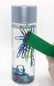 A hand using a green magnetic stick to lift colored paper clips from a clear bottle.