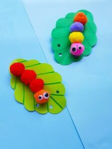 Two colorful caterpillar crafts made from pom-poms and green leaf-shaped cutouts with googly eyes, displayed on a light blue background.